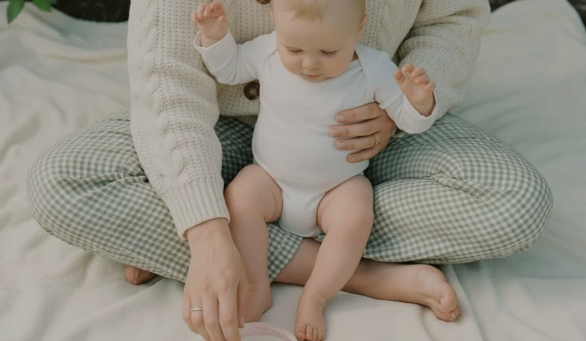 A baby in a white long-sleeve bodysuit sits on a mother's lap wearing a cream knit sweater and green-and-white gingham pants, seated on a light fabric surface, reaching for royal Heir Btanicals baby butter in Pink