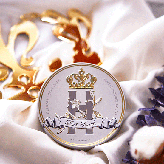 Elegant tin of Royal Heir First Touch skincare balm, featuring gold embossed details and a floral design, placed on soft white fabric with golden accents and dried botanicals.