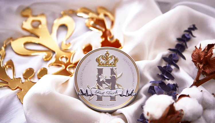 Elegant tin of Royal Heir First Touch skincare balm, featuring gold embossed details and a floral design, placed on soft white fabric with golden accents and dried botanicals.